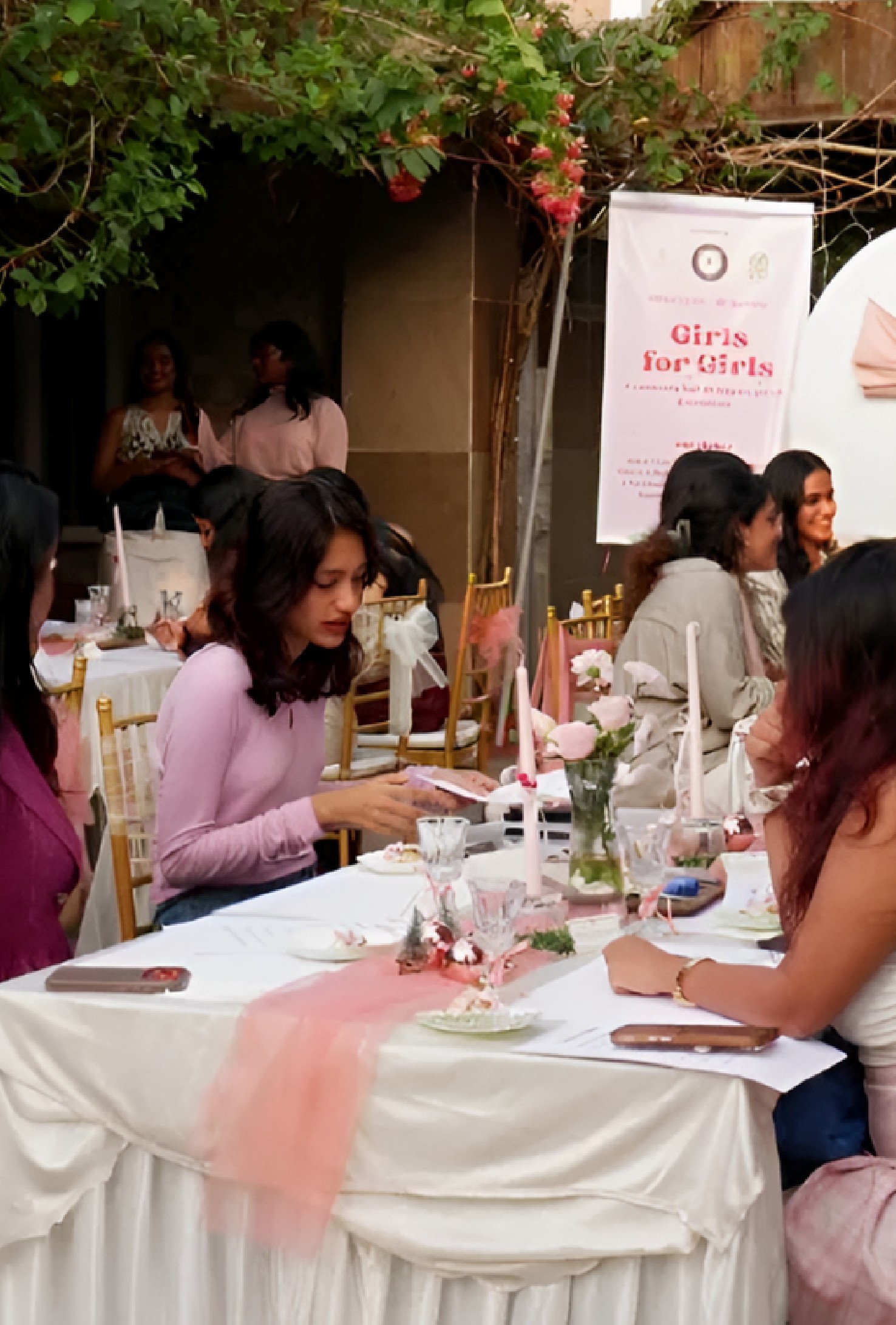 Women gathered at a pink and cream themed table setting outdoors, under a bougainvillea trellis, with a Girls for Girls Kochi banner in the background