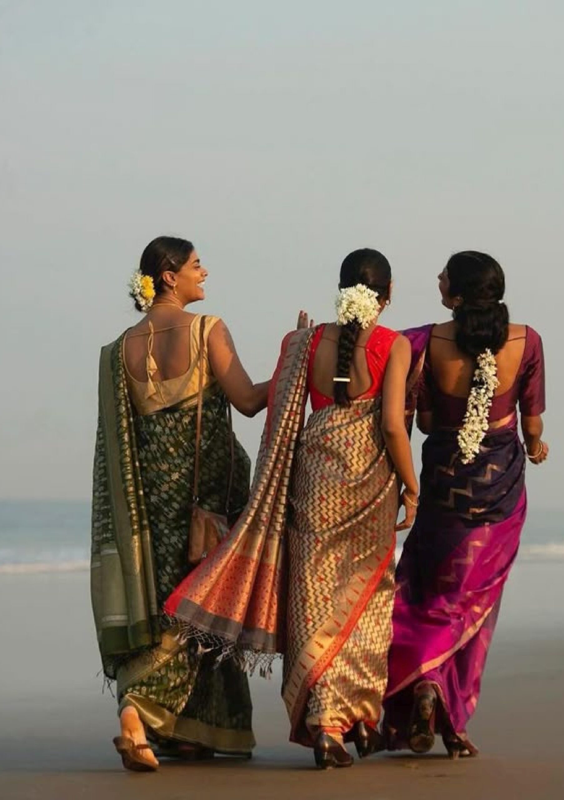 Three women in silk sarees walking together on a beach at sunset